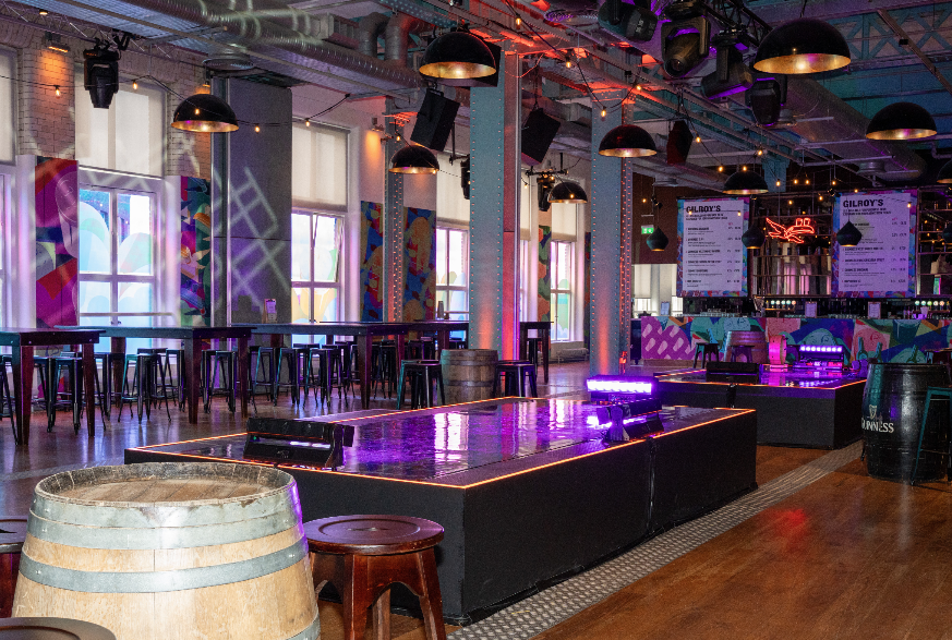 The interior of a bar with a large sleek table in the middle and a barrel in the foreground. Purple lighting reflects on the table