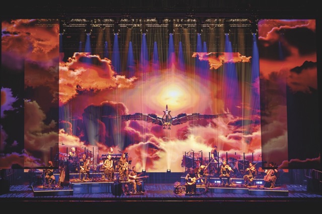 A band is set up on a stage on raised platforms. A metal dragon flies through orange and red clouds on the screen behind the stage