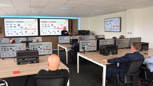 A classroom filled with adults sitting at desks with monitors in front of them. A man presents at the front with two large screens behind him