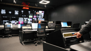 Interior of a broadcast room with rows of monitors in a blurred background and a hand pressing a button in the foreground