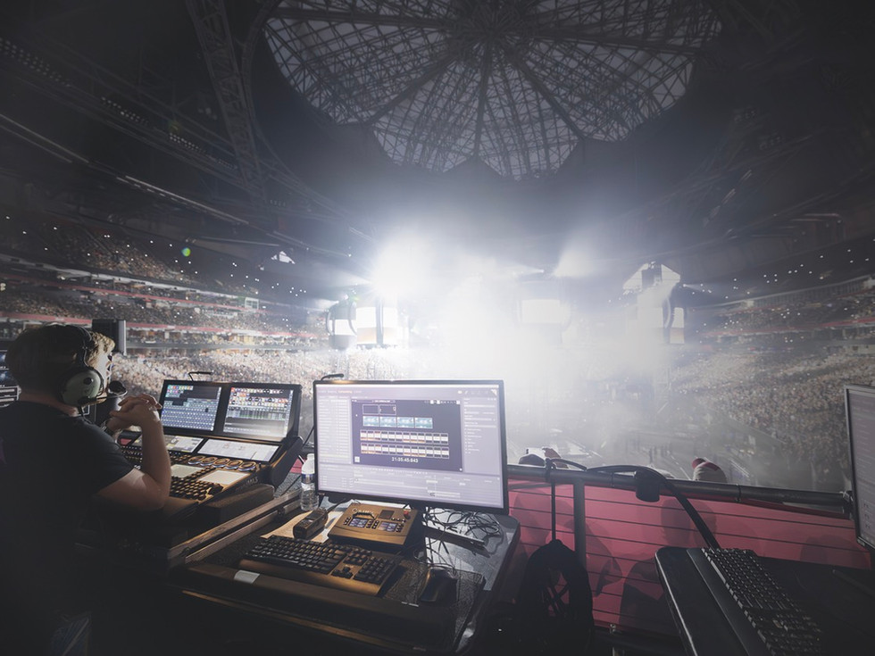 A mixing desk at the back of an arena with bright white lights at the front. A man sits at the desk working