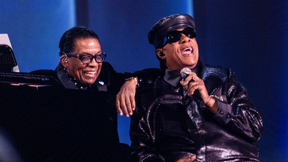 Two men sitting next to each other on stage with microphones and a blue curtain background
