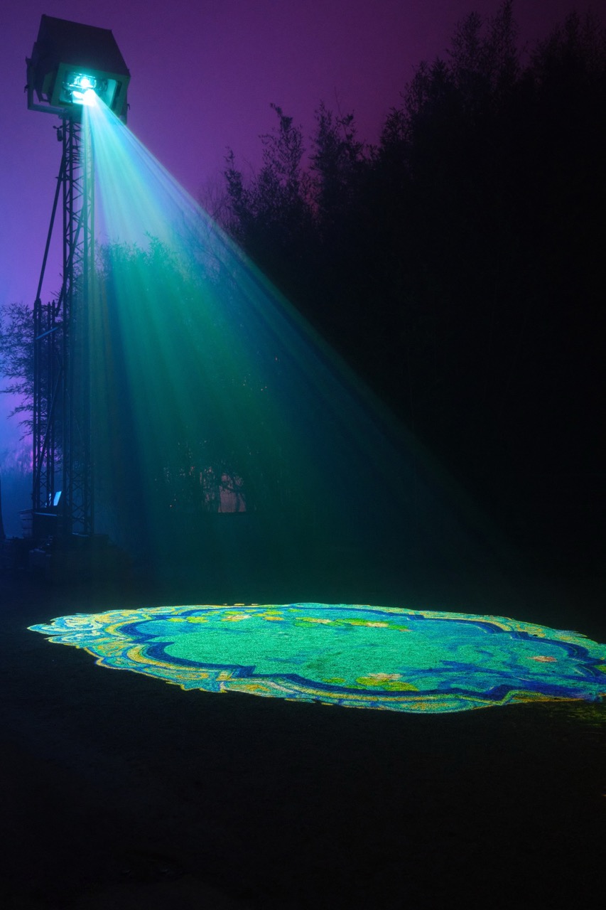 A large projector on scaffolding shine a light onto the floor making a blue pattern. The projector is placed next to a forest at night