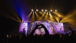 A concert with yellow stage lights and large LED screens. A crowd stands in silhouette with one person forming a heart with their hands