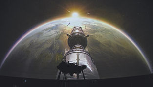A projection in a dark room showing a rocket in space facing earth with the sun behind
