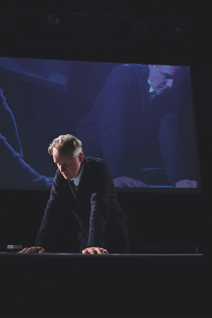 Man in dark sweater leans on table under dim lighting. A large screen behind shows the same man in a similar pose. Image Felix Mosse