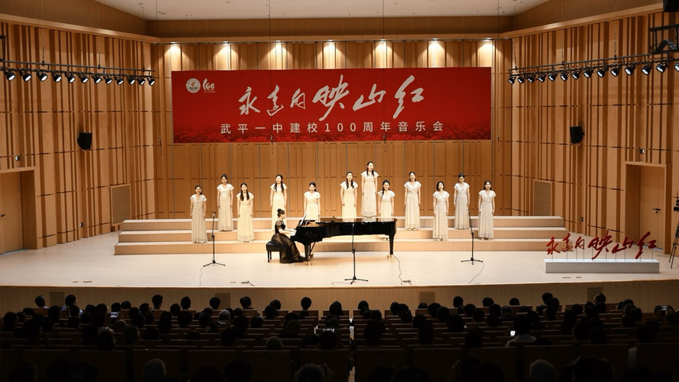 A group of women wearing white dresses sing on stage. A woman in a black dress sits at a grand piano in the centre of the stage