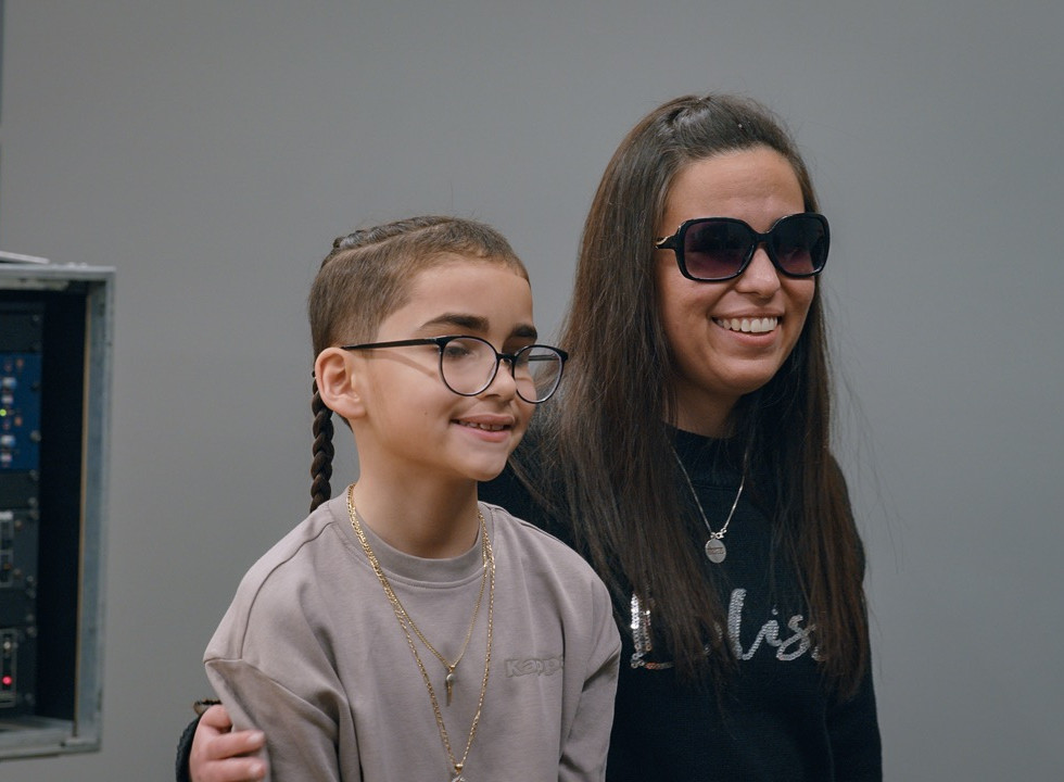 A woman with dark sunglasses sits smiling next to a young child
