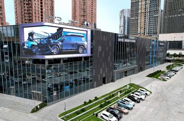 A modern, glass building with a large screen on the corner showing a car