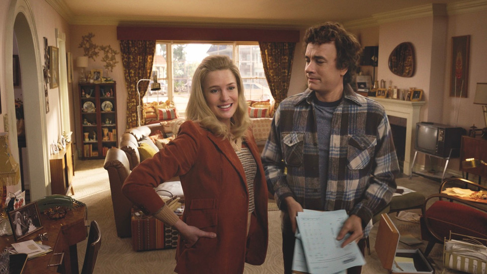 A man and a woman stand in a living room together. The man holds a stack of papers and looks at the woman. The woman smiles with her hands on her hips