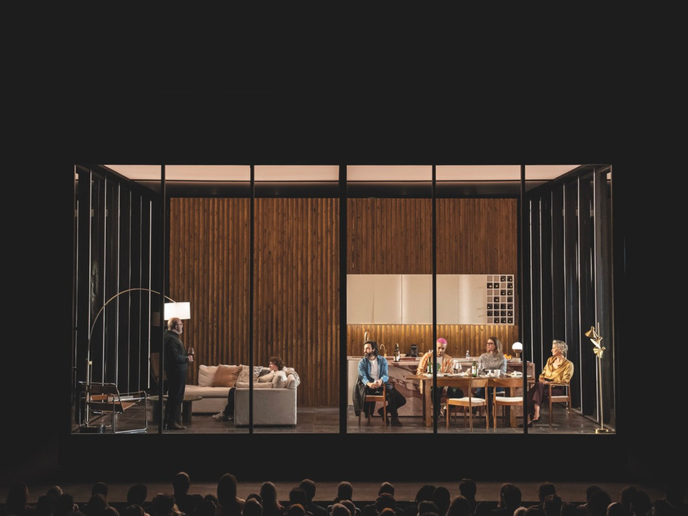 A stage show where a glass box is placed on stage with a dining room scene inside. There are actors in the box