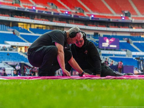 Two men in black t-shirts and headphones working on a pink blanket on grass in stadium