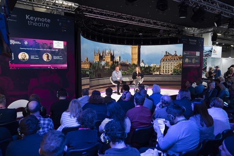 A man and a woman sitting on a stage delivering a presentation to a crowd lit in blue light. Behind the pair, there is an LED screen showing London