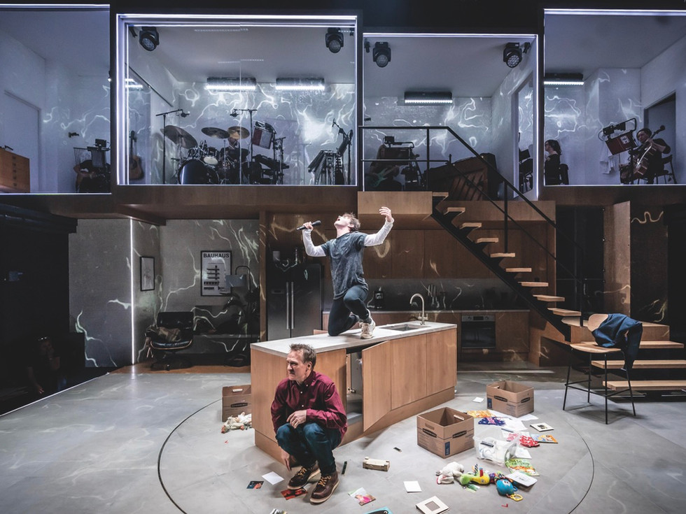 Two actors stand on a stage. One is crouched on the messy floor while the other roars on a counter. There are instruments placed in glass boxes at the back of the stage