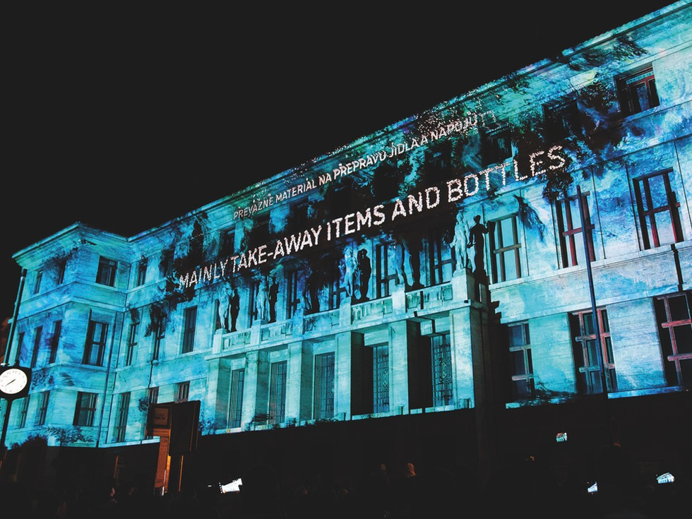 Building facade at night lit with projected blue light. Text reads "MAINLY TAKE-AWAY ITEMS AND BOTTLES." Dark sky backdrop.