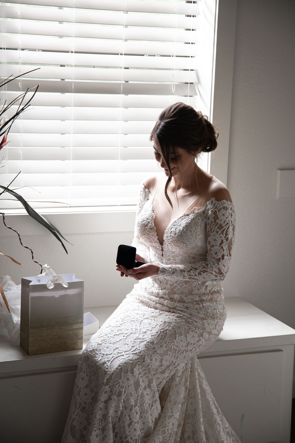Bride in a lace wedding dress sitting by a window holding a ring box