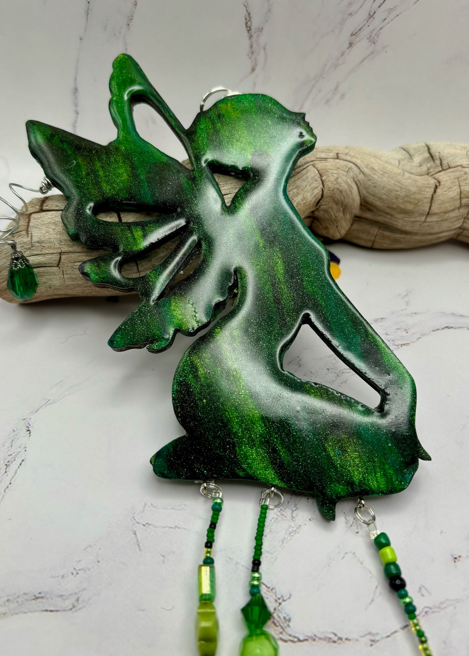 Green fairy figurine with wings and dangling beads next to driftwood background.