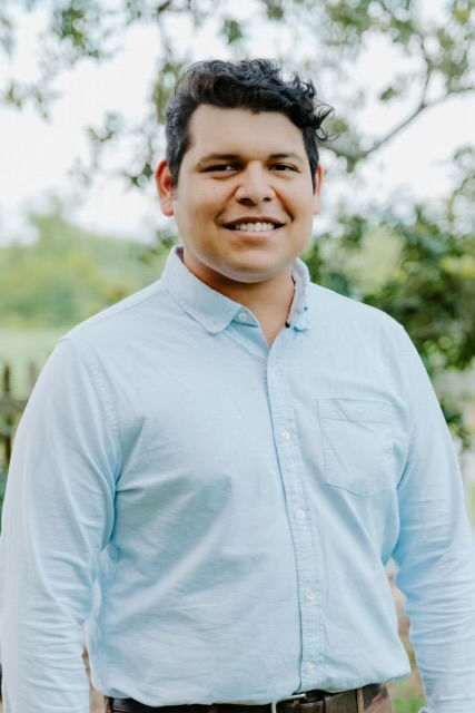 Smiling person in a light blue shirt stands outdoors with green trees in the background; a relaxed, cheerful atmosphere is conveyed.
