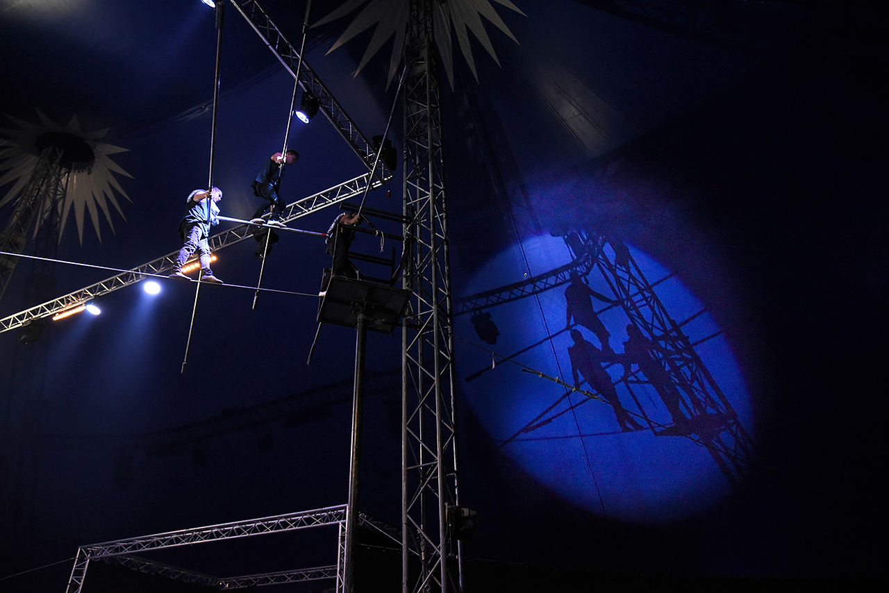 Acrobates sur trapèze du Cirque Infernal sous projecteur bleu.