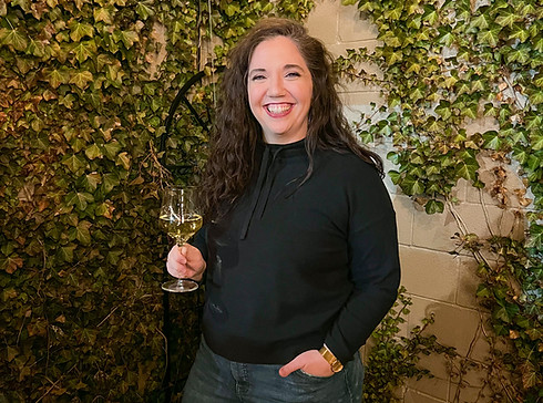 A woman is standing in front of an ivy covered wall holding a wine glass and smiling.