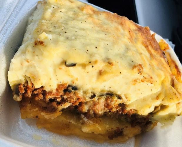 A food takeout container is filled with a large piece of Greek moussaka filled with meat and covered with a creamy cheese sauce