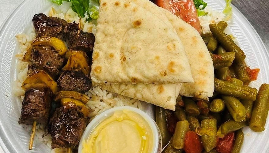 A plate of beef kebabs, rice, green beans, Greek salad, hummus and pita