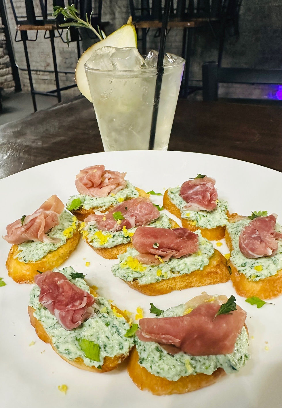 Goat cheese toasts topped with prosciutto fill a plate with a pear rosemary cocktail on the side.