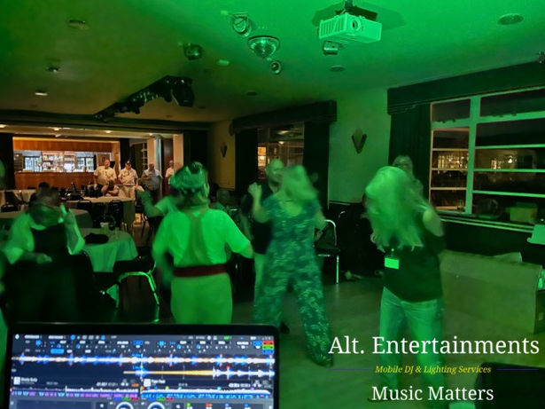 Guests dancing energetically at a disco event at Sywell Aerodrome in Northamptonshire, organized for The Classic Corvette Club. The photo, taken by Alt. Entertainments—a Mobile DJ, Disco, and Events Company—captures a vibrant dance floor illuminated by colorful disco lights, showcasing the lively atmosphere and enjoyment of the attendees.
