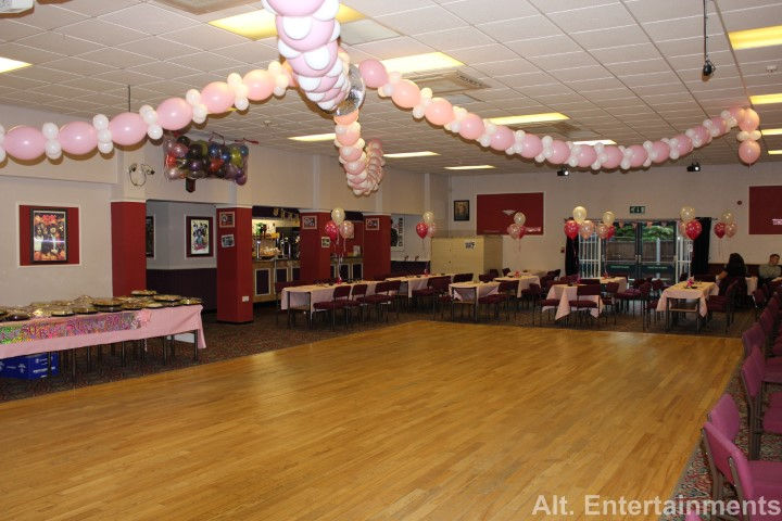 Photograph of the function room at the West End Club in Merridale, Wolverhampton, prepared by Alt. Entertainments, a mobile DJ, disco, and events company. The spacious room features elegantly arranged tables with white linens, stylish chair covers, and matching tableware. Floral centerpieces add a touch of natural beauty. Ambient lighting creates a warm glow, highlighting the seating and dance floor areas. State-of-the-art sound and lighting equipment is discreetly positioned, ready for high-quality entertainment. The setup is versatile, accommodating both intimate gatherings and larger parties, reflecting the company's dedication to client satisfaction.