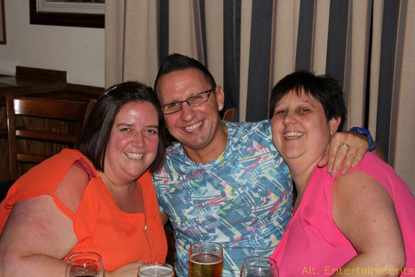 A group of people joyfully poses for a photo at Tettenhall Albert Lawn Tennis Club in Wolverhampton. The guests, dressed in elegant and stylish outfits, stand closely together with expressions of happiness and camaraderie. The lush greenery and well-maintained tennis courts in the background add to the elegance of the scene. A professional DJ setup by Alt. Entertainments is visible, contributing to the lively atmosphere of the event.