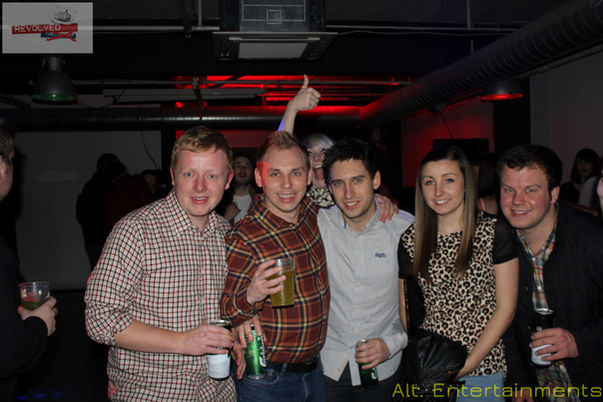 A lively group of people posing for a photo at the Revolved Indie Night Reunion, held at the Fixxion Warehouse in Wolverhampton. The venue, which used to be the Light Bar, provides a modern, industrial backdrop for the indie-themed event. The photo, taken by Alt. Entertainments, showcases the attendees' fashionable indie attire and the vibrant atmosphere of the event.