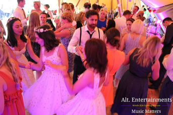 A lively scene of wedding guests dancing at a reception held at Market Overton, Oakham, Rutland. The dance floor is illuminated by soft, romantic lighting that enhances the festive atmosphere of the celebration. Guests of all ages, dressed in their wedding best, are seen enjoying the music, which is provided by Alt. Entertainments, a Wedding DJ, Mobile DJ, Disco & Events company. The DJ setup is visible in the background, creating the perfect blend of music for the joyous occasion. Smiling and laughter fill the room as guests celebrate the wedding in high spirits.