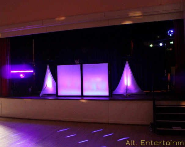 A professional mobile DJ setup by Alt. Entertainments at Middlewich Civic Centre, Cheshire. The setup includes a modern DJ booth with advanced sound equipment, surrounded by vibrant LED lighting that creates a dynamic and festive atmosphere. The lighting and sound elements are arranged to enhance the party experience, with the booth’s sleek design contributing to a visually impressive display. The setup is perfect for high-energy events, with a focus on delivering quality entertainment.