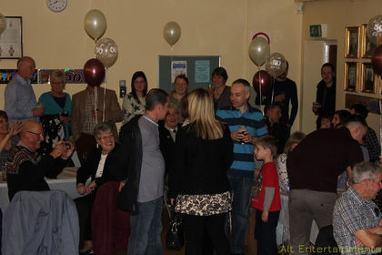 A lively crowd enjoys a party at Braunstone Civic Centre in Leicester, Leicestershire. Partygoers are seen dancing, laughing, and chatting, immersed in the festive atmosphere created by Alt. Entertainments, a mobile DJ, disco, and events company. The venue is decorated with vibrant colors, balloons, and streamers, with professional lighting enhancing the celebratory mood.
