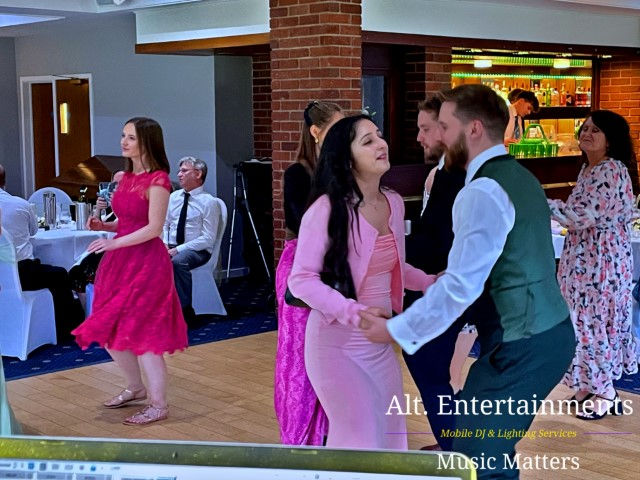 Wedding guests dancing enthusiastically at a reception held at Hagley Golf Club in Stourbridge. The scene depicts a lively dance floor with guests in formal attire actively participating in the celebration. In the background, a well-equipped DJ setup from Alt. Entertainments is visible, featuring colorful lighting and professional sound equipment, adding to the festive mood of the event.
