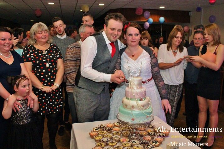 Bride and groom happily cutting their wedding cake at Fordhouses Cricket Club in Wolverhampton. The multi-tiered cake is beautifully decorated with floral designs. Guests surround the couple, sharing in their joy. Captured by Alt. Entertainments, the scene exudes love, celebration, and meticulous wedding planning.