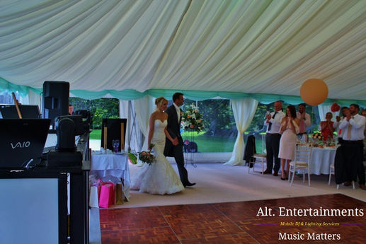 Bride and groom making their grand entrance at a marquee wedding in Aberystwyth, Wales. The photo, taken by Alt. Entertainments, shows the couple walking into the marquee surrounded by beautifully decorated tables and elegant lighting. The bride is in a stunning gown, and the groom is in formal attire. The image captures the festive and romantic atmosphere of the wedding celebration, highlighting the meticulous setup and decoration of the venue.