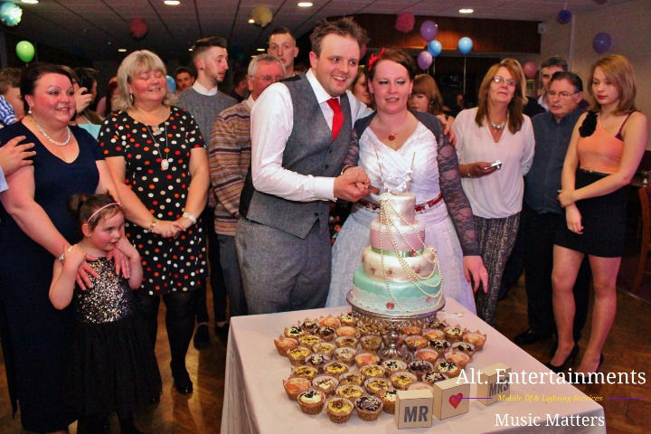 Bride and groom joyfully cutting their beautifully decorated wedding cake at Fordhouses Cricket Club in Wolverhampton. The multi-tiered cake features intricate floral designs. Guests surround the couple, smiling and applauding. Photo taken by Alt. Entertainments, highlighting the love, celebration, and expert event planning of the day.