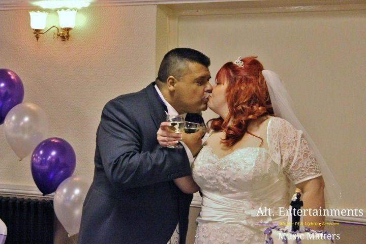 The bride and groom pose for a wedding photo in Bilston, Wolverhampton. The bride is in a white gown with lace and beadwork, standing beside the groom in a tailored suit. They are surrounded by vibrant floral arrangements and lush greenery. The photo, taken by Alt. Entertainments, captures their joy and love on their special day.