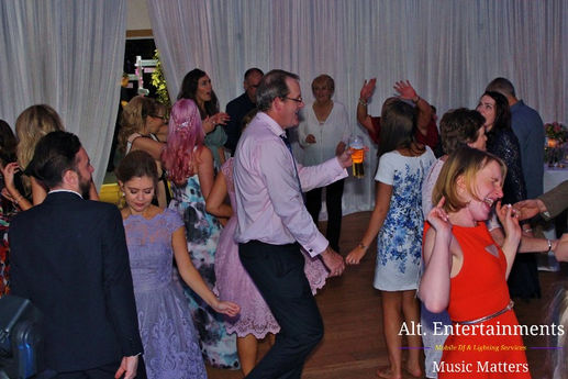 A vibrant crowd of wedding guests dance energetically at a disco in Crosby, Liverpool. Captured by Alt. Entertainments, the photo highlights a packed dance floor filled with smiles and laughter, with colorful lighting enhancing the joyful atmosphere. The image reflects the DJ’s expert ability to keep the party alive, making the night an unforgettable celebration.