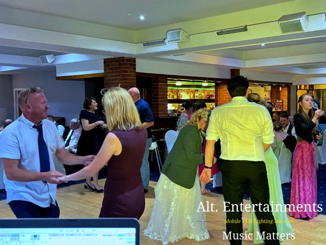 Wedding guests dancing energetically at a reception held at Hagley Golf Club, Stourbridge. The scene captures guests enjoying the lively atmosphere, illuminated by festive lighting. The image, taken by Alt. Entertainments, highlights their expertise in providing mobile DJ, wedding DJ, and event services, showcasing their contribution to a vibrant and engaging event experience.