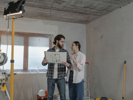 A male and female standing inside a house ongoing repair, discussing about a blueprint.
