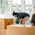 A female writing on a box, while a male is arranging in the background, surrounded by house moving boxes.
