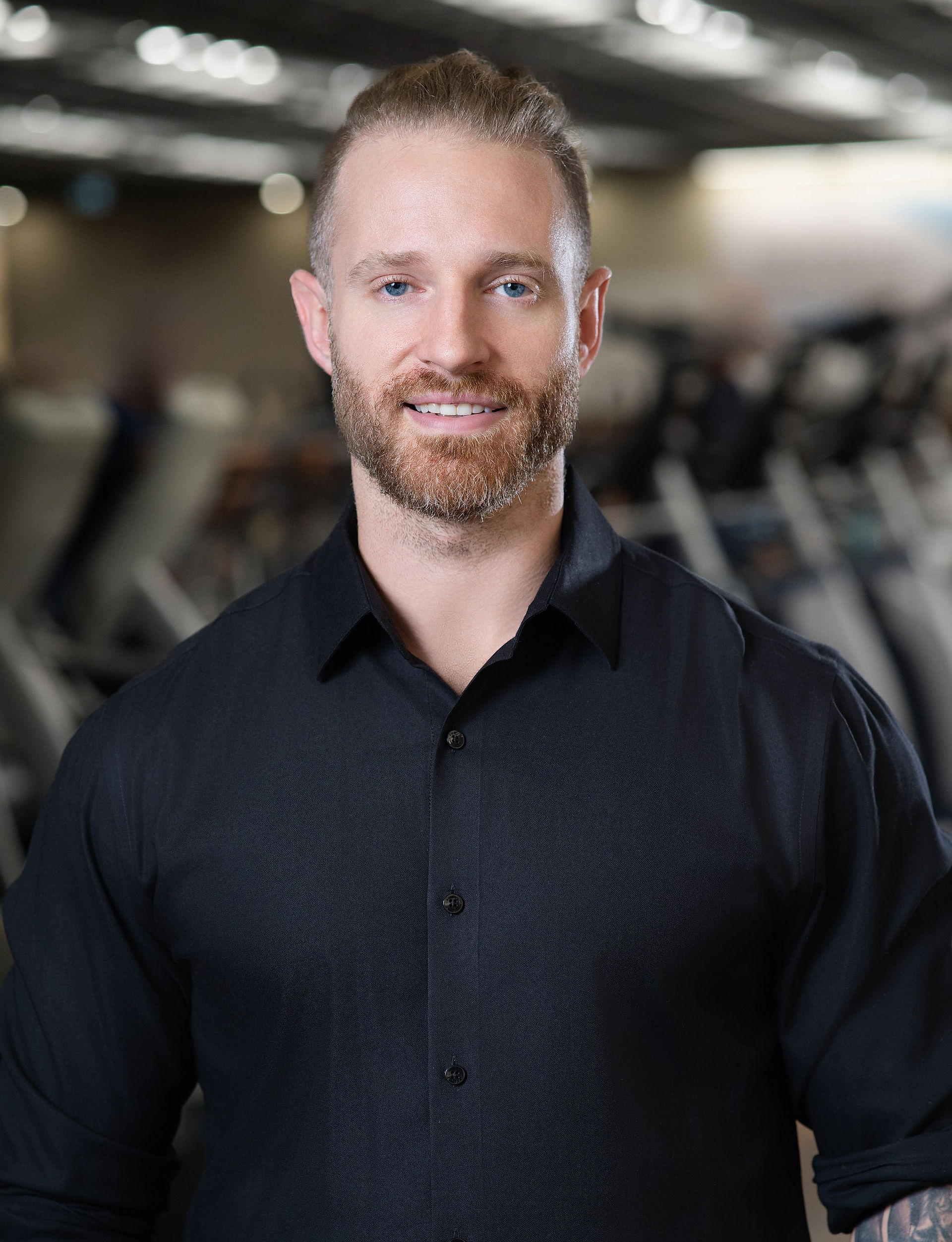 Professional Headshot Movati executive wearing black shirt with gym in background.
