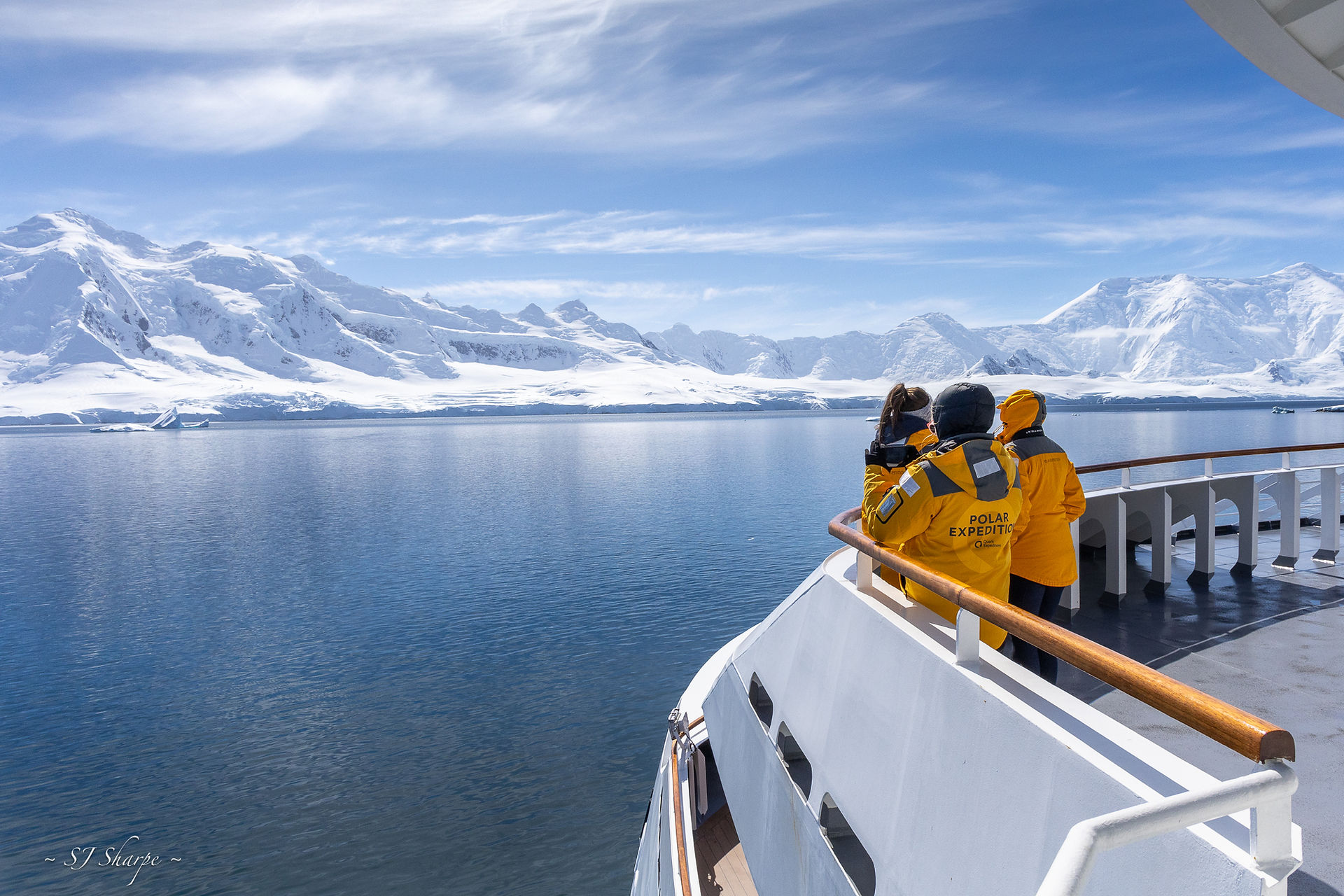 A small group of Antarctica travellers stand on the viewing deck of an expedition cruise ship, enjoying the views of beautiful, snow-covered mountains