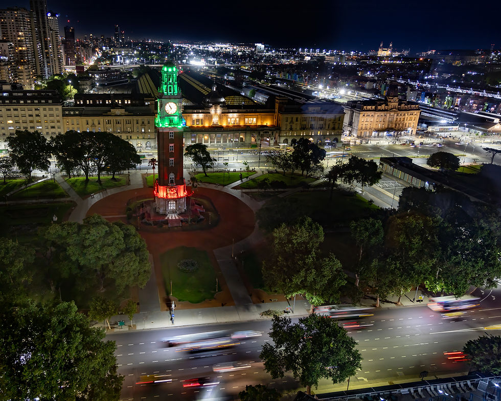 Illuminated Torre Monumental clock tower with green and red lights stands in a circular plaza at night, surrounded by trees and Buenos Aires city lights in the background
