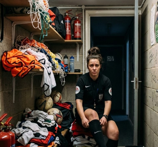 Woman football referee sits in a cramped locker room, adjusting a black sock beside piles of football kits, balls, and fire extinguishers.