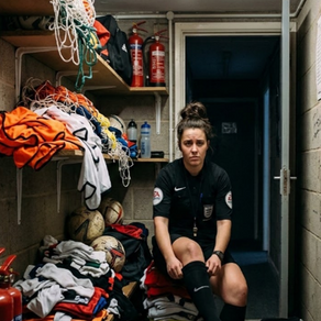 Woman football official sits in a cramped locker room, adjusting a black sock beside piles of football kits, balls, and fire extinguishers.
