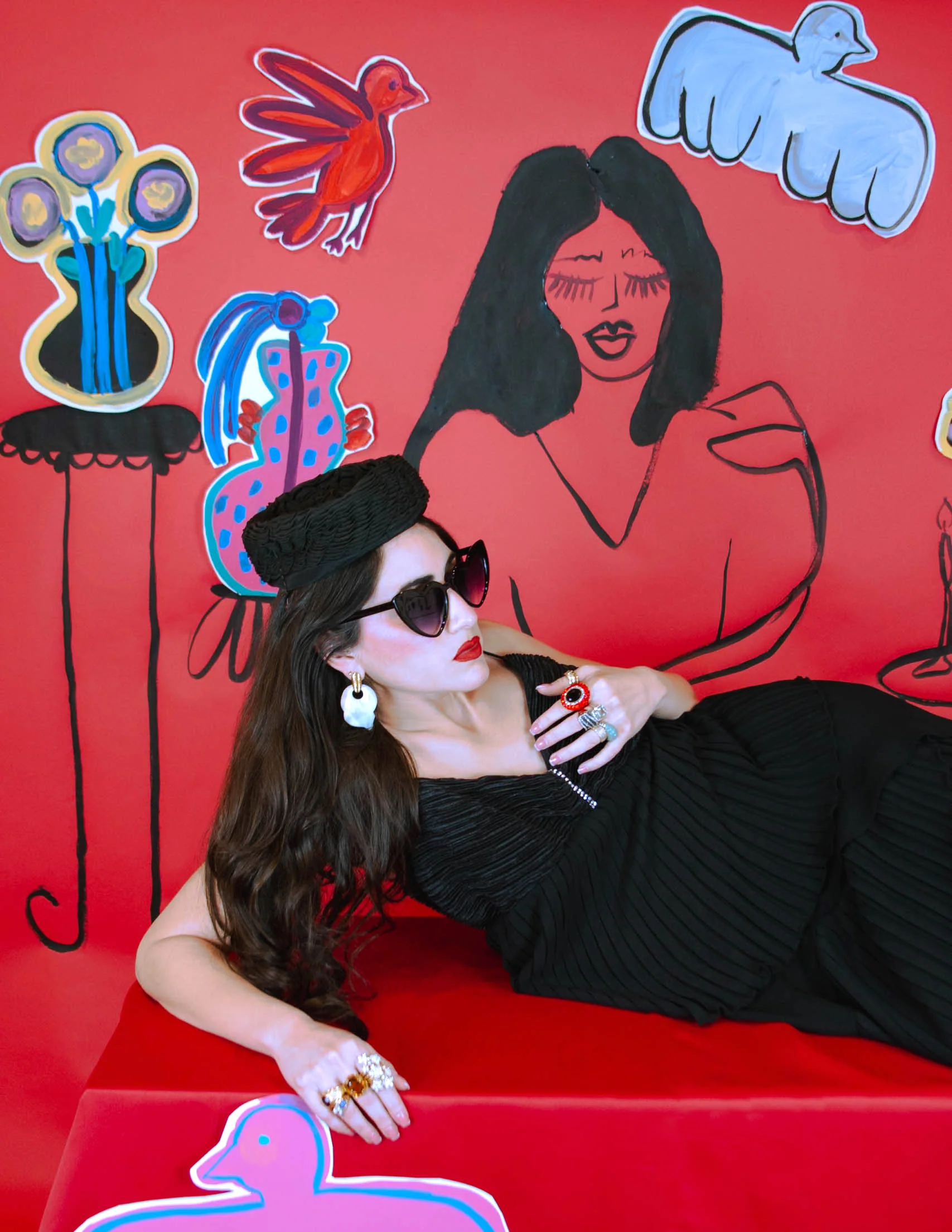 Persona con vestido negro, sombrero y gafas de sol recostada sobre superficie roja, con dibujos artísticos de pájaros y flores en el fondo.