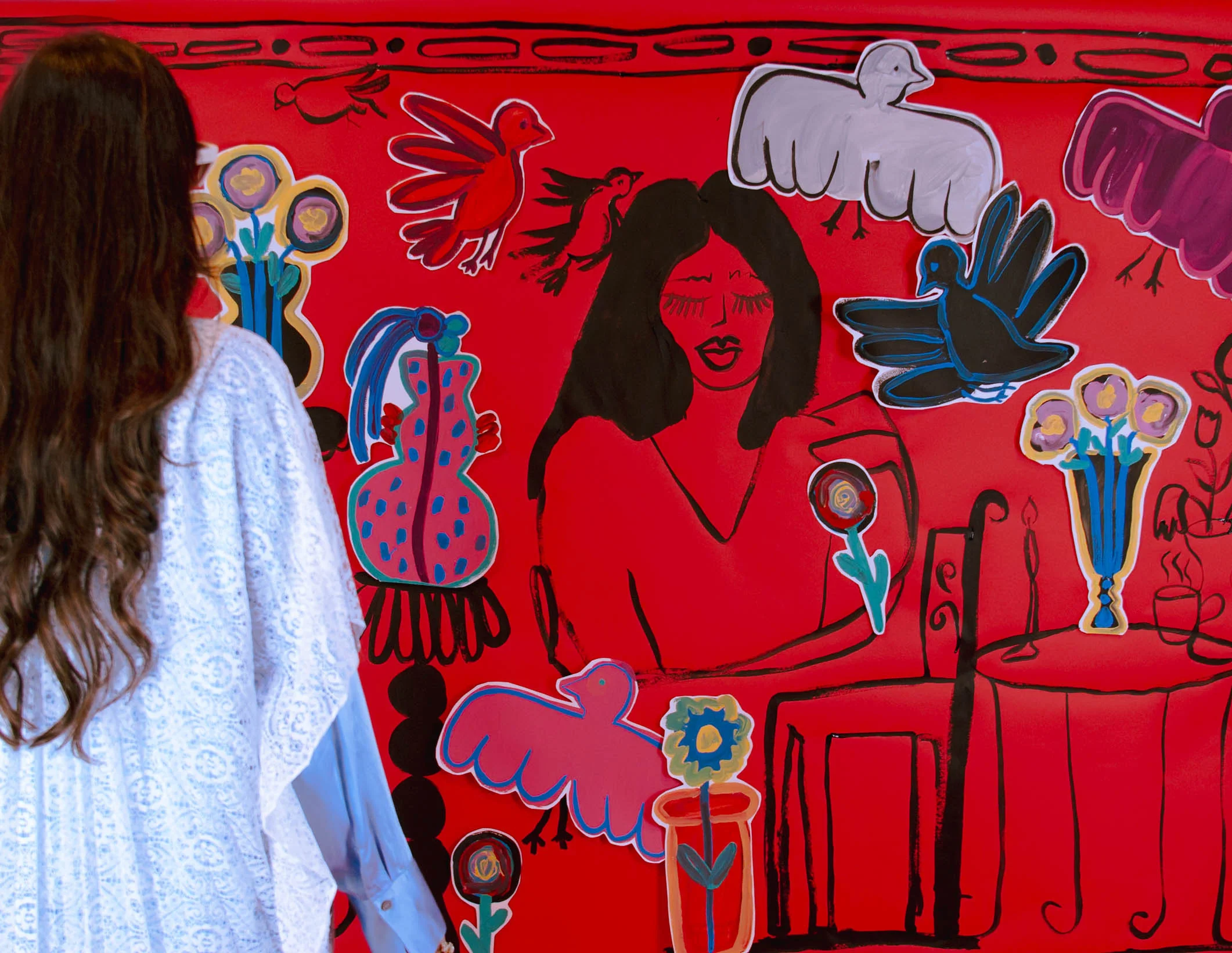 Vista lateral de una persona con cabello largo y oscuro vistiendo un conjunto blanco de encaje, observando un mural. El mural sobre fondo rojo contiene ilustraciones de aves coloridas, flores, y una figura femenina dibujada con cabello negro. La decoración incluye varios elementos ilustrados como mesas y floreros.